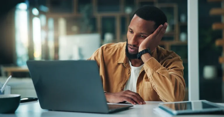Programador cansado con la cabeza apoyada en la mano frente al portátil, como quien espera a que se revise el código que escriben los agentes de IA