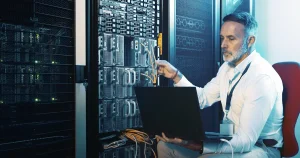 Un técnico revisa el cableado de un armario de servidores con un portátil sobre las rodillas, el tipo de sistemas críticos que la Cyber Mission de Anthropic quiere ayudar a proteger