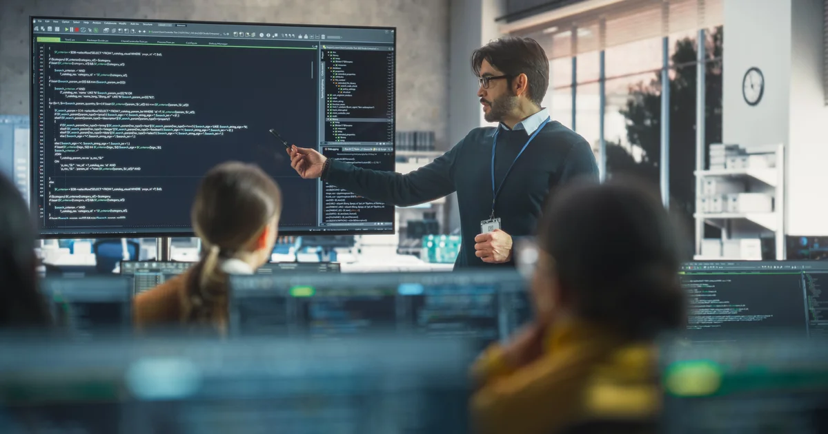 Un ingeniero señala código en una gran pantalla mientras sus compañeros siguen desde sus puestos una formación sobre cómo llevar la IA a una empresa
