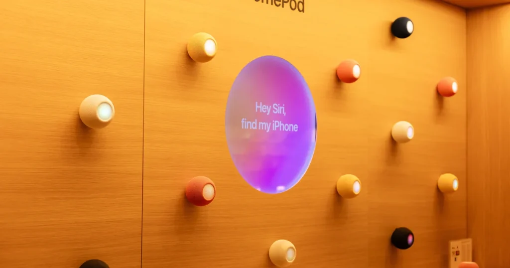 Altavoces HomePod mini de varios colores en una pared de una Apple Store, alrededor de una pantalla que pide a Siri encontrar un iPhone, en la noticia sobre la nueva Siri AI de Apple para casa
