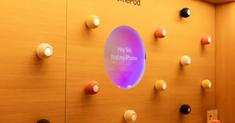Altavoces HomePod mini de varios colores en una pared de una Apple Store, alrededor de una pantalla que pide a Siri encontrar un iPhone, en la noticia sobre la nueva Siri AI de Apple para casa