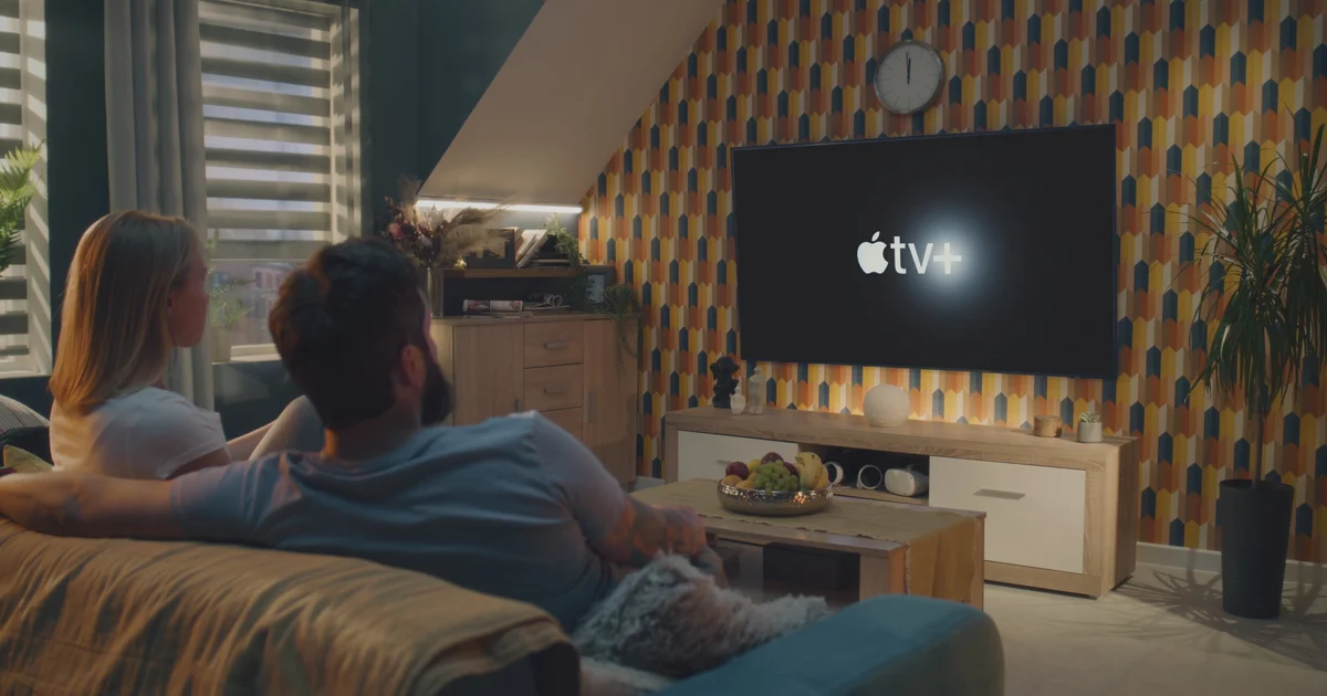 Una pareja en el sofá viendo una televisión de pared con el logotipo de Apple TV+, el tipo de salón donde encaja el nuevo Apple TV 4K que sí se vende en España