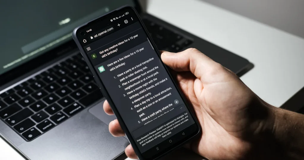 Una mano sostiene un móvil con una conversación de ChatGPT abierta delante de un portátil, la app donde OpenAI probará anuncios visuales