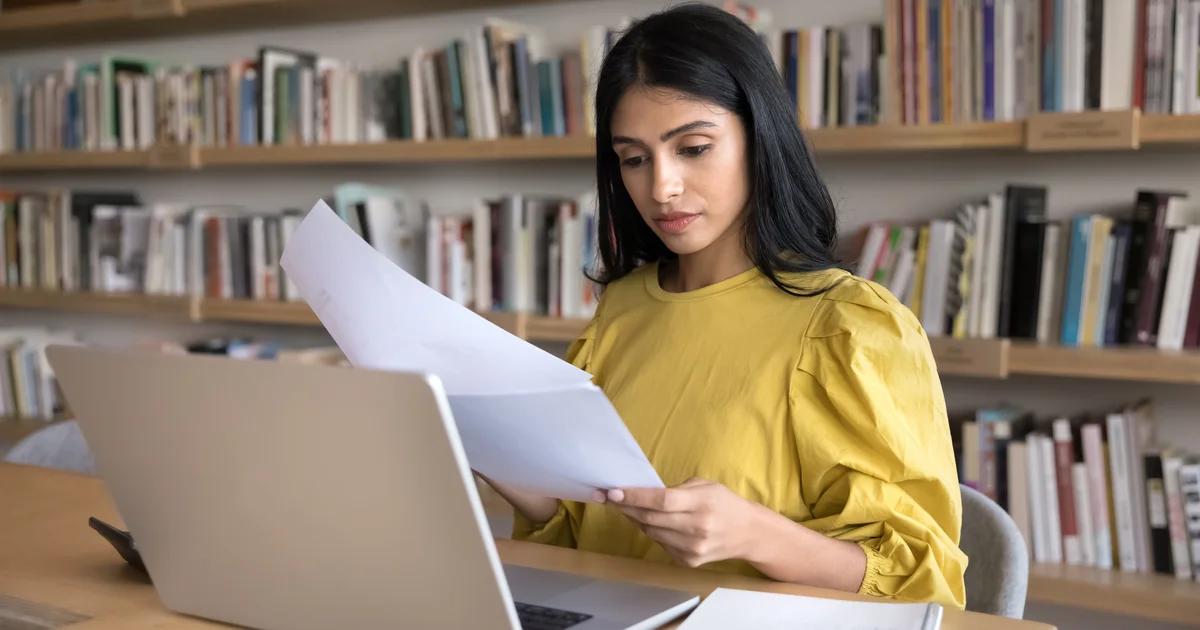 Mujer ante un portátil leyendo unos folios impresos en una biblioteca, como quien repasa la letra pequeña de las nuevas normas de uso de Claude