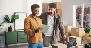 Dos emprendedores revisan una tableta en su pequeño negocio, rodeados de cajas de envío, para ilustrar las nuevas herramientas de Gemini para pymes