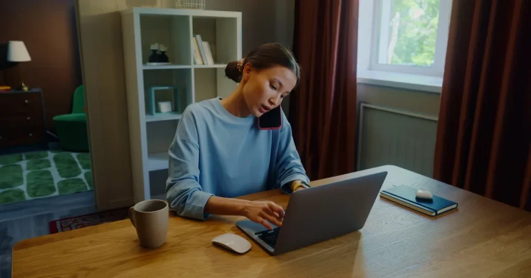 Mujer hablando por el móvil mientras escribe en un portátil en el escritorio de casa, el tipo de trabajo de oficina y programación al que apunta Kimi K3.1 Pro